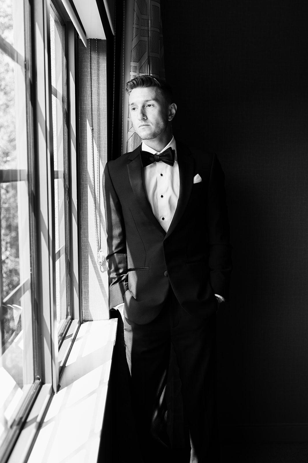 Black and white portrait of the groom standing by a window with hands in his pockets, looking thoughtful.